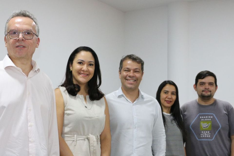 Cinco pessoas posam sorrindo para a foto em um ambiente interno de paredes brancas. Dois homens vestem camisa social clara, uma mulher usa vestido bege, outra mulher está de camiseta listrada e calça jeans, e um homem veste camiseta cinza com estampa colorida.